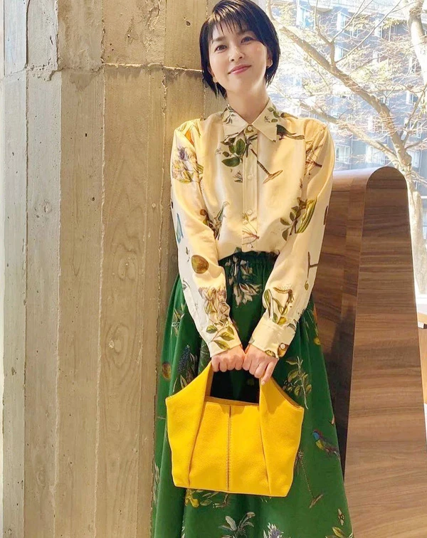 松隆子很適合這種帶點文藝感的穿搭。她用植物圖鑑印花襯衫搭配森林綠 A 字長裙，雖然上下身都有印花元素，但因為整體色調都落在綠色、米色和黃色這種偏自然的大地色系，所以看起來完全不會太花，反而有種很舒服的層次感。她把襯衫下擺紮進裙子裡，也讓整體比例變得更俐落、有精神。最亮眼的還是那款鮮黃Tod's色托特包，剛好替整體偏沉穩的配色多了一點活力，讓這套穿搭看起來既有氣質，又不會太成熟。