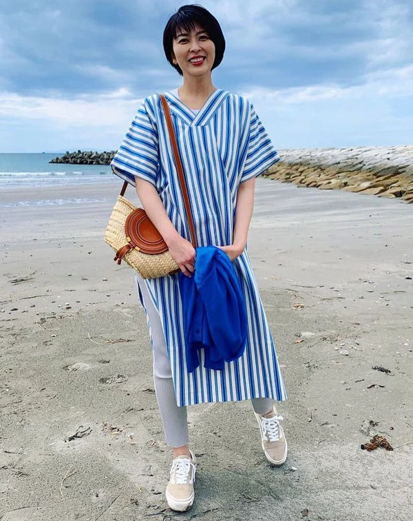 站在海邊的松隆子，選擇了最能代表清爽感的海軍藍與白色交錯的直條紋寬鬆長洋裝，側邊開衩的設計，隱約露出內搭的淺灰色羅紋緊身褲與休閒帆布鞋。「藍、白配」，可說是視覺減齡法當中，最萬年不敗的黃金公式。這是，只要手上隨性抓著同色系的藍色針織外套，再斜背一個皮革拼接草編包，便能瞬間注入滿滿的空氣感與悠閒少女氣息，完全打破我們對於50歲就是長者的沉悶感喔！