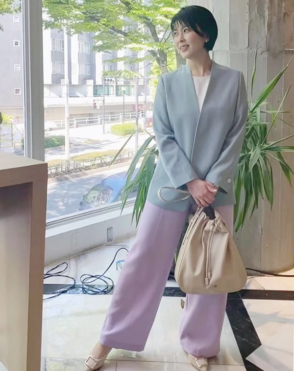 誰說過50歲就不能穿彩色？這套搭配絕對是熟齡女性一定要學起來的「高階色彩減齡術」！  照片中，松隆子打破了職場正裝非黑即白的沉悶框架，大膽將兩種柔和的馬卡龍色系疊加。無領設計的灰藍色西裝外套，內搭純白上衣，下身則大膽配上一條薰衣草紫的寬褲。