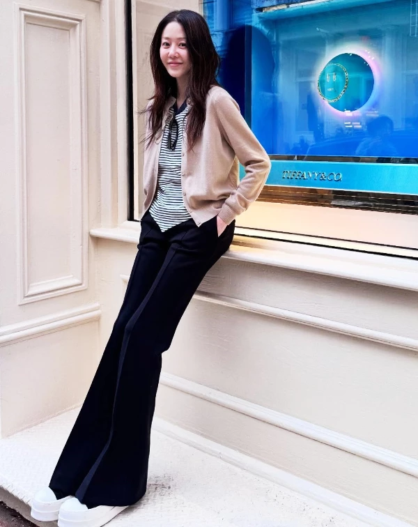 高賢廷倚靠在Tiffany珠寶櫥窗前的私服照，完美詮釋了何謂「優雅的鬆弛感」，那抹淡雅、從容的笑容，完全呈現一種與歲月共行的自在氣息。至於穿搭上，高賢廷聰明地以溫柔的燕麥色針織衫，疊搭度假氣息的條紋內搭，並利用柔和的中性色調來提亮整體氣色，精準平衡溫潤與幹練；下半身，則是選用高腰的小喇叭寬褲，並搭配厚底小白鞋，不僅巧妙延伸長腿比例，更為整體造型注入如女大生般的清爽活力。