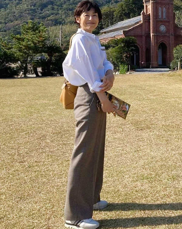 在藍天綠地襯托下，鈴木保奈美以寬鬆的廓形白襯衫，搭配格紋高腰寬褲與運動鞋，後背一個焦糖色皮革包，整個人顯得非常輕盈自在。一般人在穿基本款白襯衫時，千萬不要把釦子扣得死緊，或選太合身的版型。學她挑選落肩、帶有空氣感的寬鬆襯衫，將袖口隨意挽起，並把下擺紮進高腰褲中帶出好比例，配上一雙厚底白鞋，就能完美複製這份兼具舒適與文藝感的旅行逆齡感。