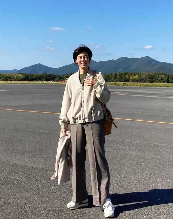站在遼闊停機坪上的鈴木保奈美，換上了極具空氣感的米白色立領休閒夾克，下半身則混搭了一條垂墜感極佳的棕色格紋寬褲與乾淨白鞋。一般人穿運動風夾克時，很容易顯得太邋遢或像去運動，祕訣就在於「風格混搭」。學她用帶有正裝感的細格紋西褲來搭配輕便的廓形夾克，內搭則選擇低調的素色T，這種「半休閒、半正式」的組合，不僅穿起來舒適，更能展現出不經意、放鬆，且極度減齡的文藝旅人風範。