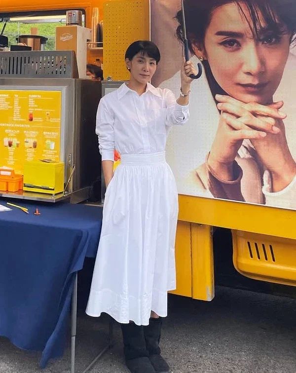同樣是在應援餐車前，金瑞亨卻展現了少見的全白知性造型。上身是乾淨的立領白襯衫，下身搭配一條帶有細緻抓褶設計的高腰白色A字長裙，腳下則踩著一雙黑色休閒短靴。全白色調，不僅在視覺上極具延伸感，更在隨性中注入了一抹優雅的文藝氛圍。