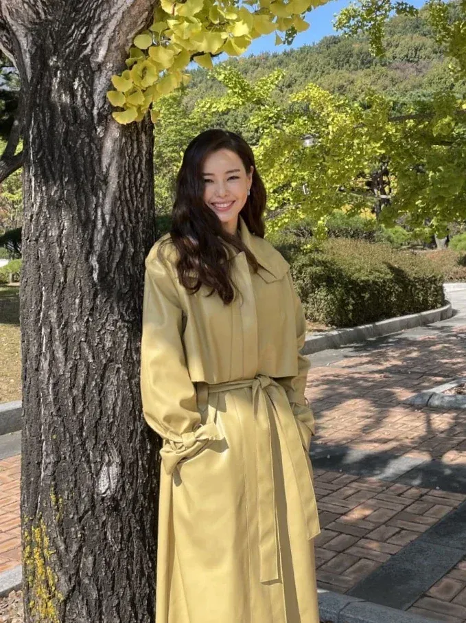 皮革風衣,往往容易給人沉重或過於銳利的霸氣感,但李荷妮聰明地挑選了帶著淡淡奶油黃的皮革材質。在陽光與銀杏樹下,這種柔和的淺亮色調能自然為肌膚打上「嫩膚濾鏡」,既保留了風衣特有的率性俐落,又完全消解了熟齡感常見的嚴肅僵硬。
