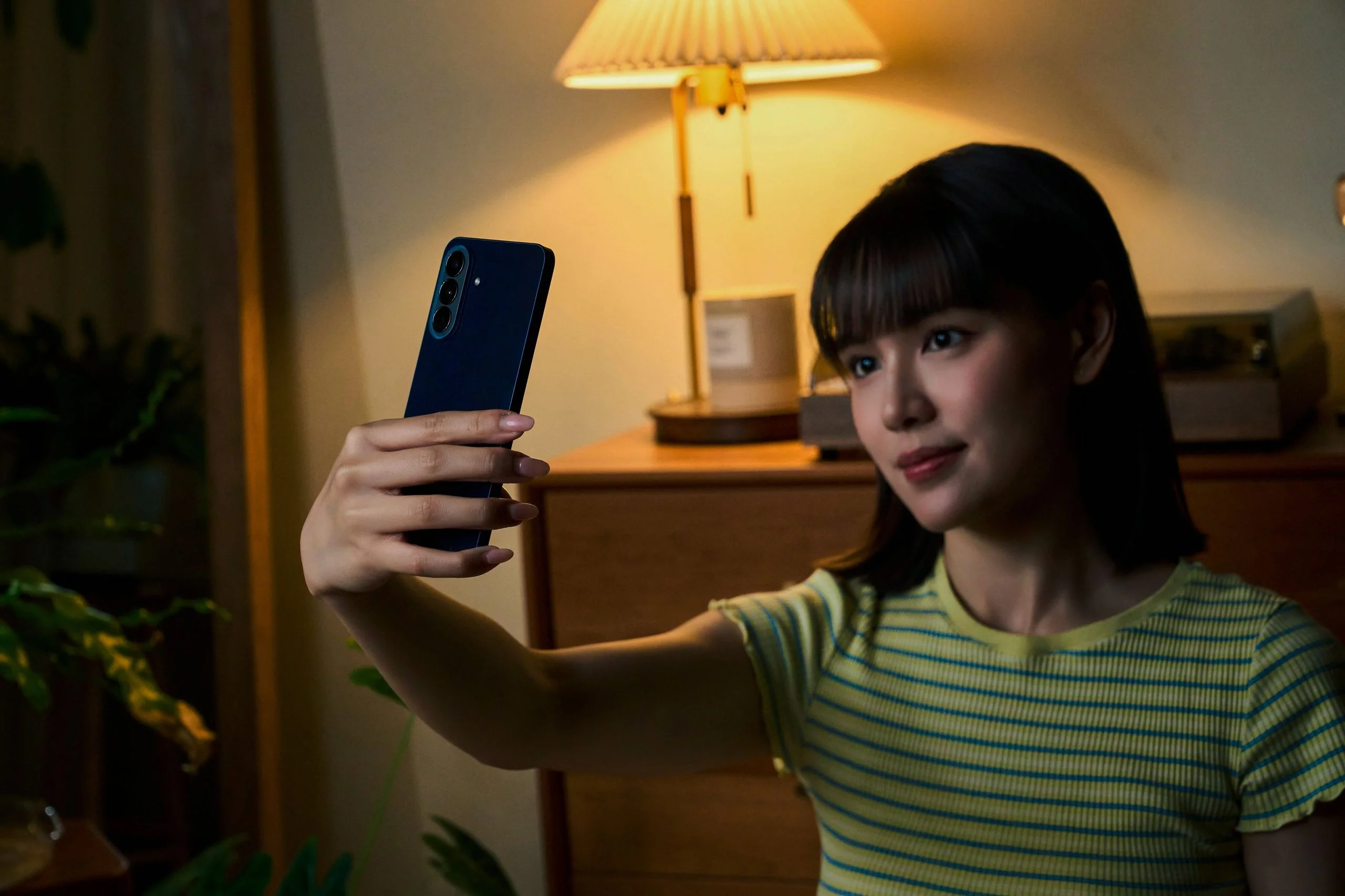 Model 拿著雅痞藍 Galaxy A57 5G 於昏暗室內自拍