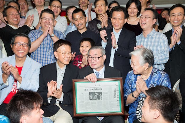 發叔獲頒大學名譽院士後，歷屆幹事為他舉辦一個大型慶祝晚會，發叔與發嬸上台與嘉賓合影。發叔拿着《人民日報》的報道文章紀念相架。（灼見名家圖片）